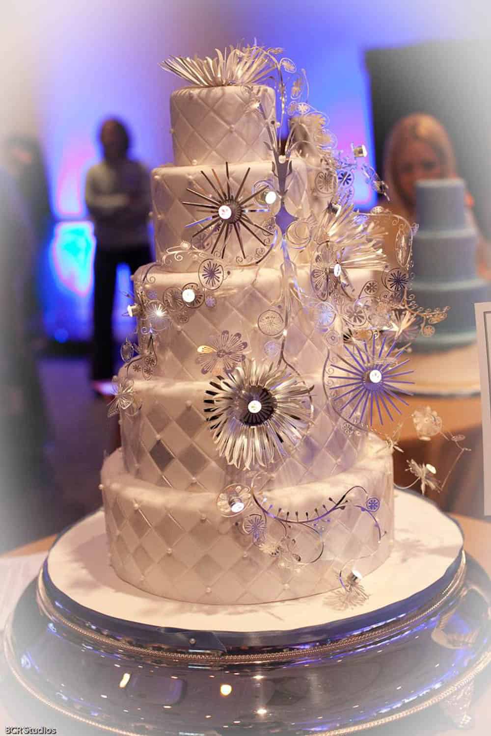A multi-tiered white wedding cake with quilted patterns and decorated with silver, floral, and jeweled embellishments sits on a round table.