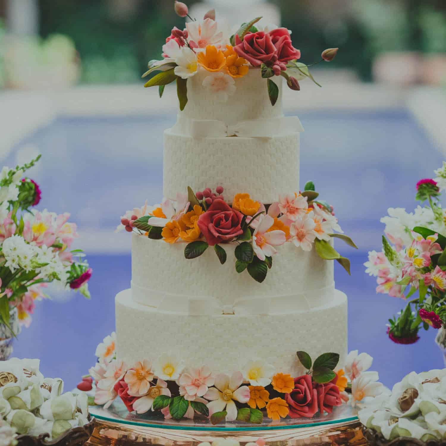 Three-tiered white cake decorated with red, yellow, and pink flowers, displayed on a table with floral arrangements in the background.