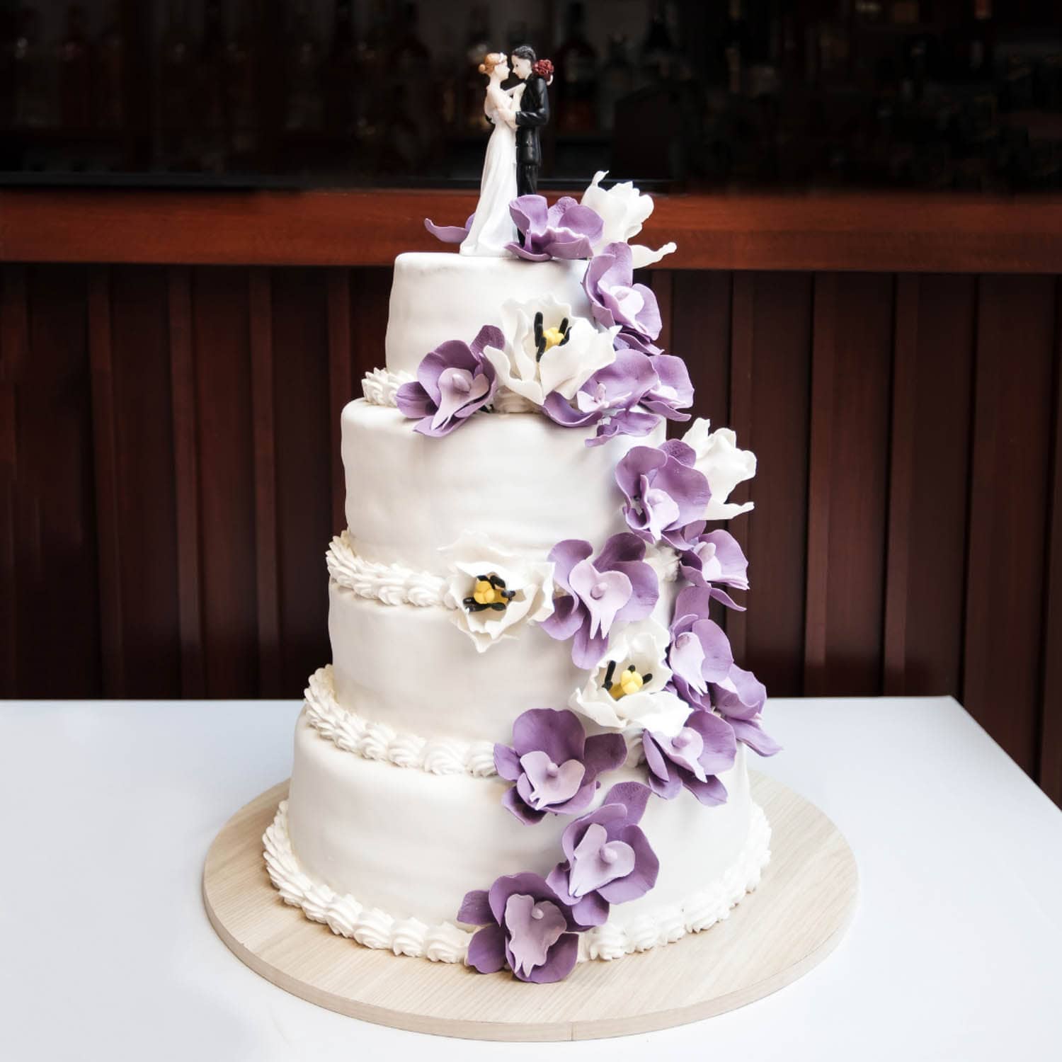 Three-tiered white wedding cake decorated with purple and white flowers, topped with a bride and groom figurine.