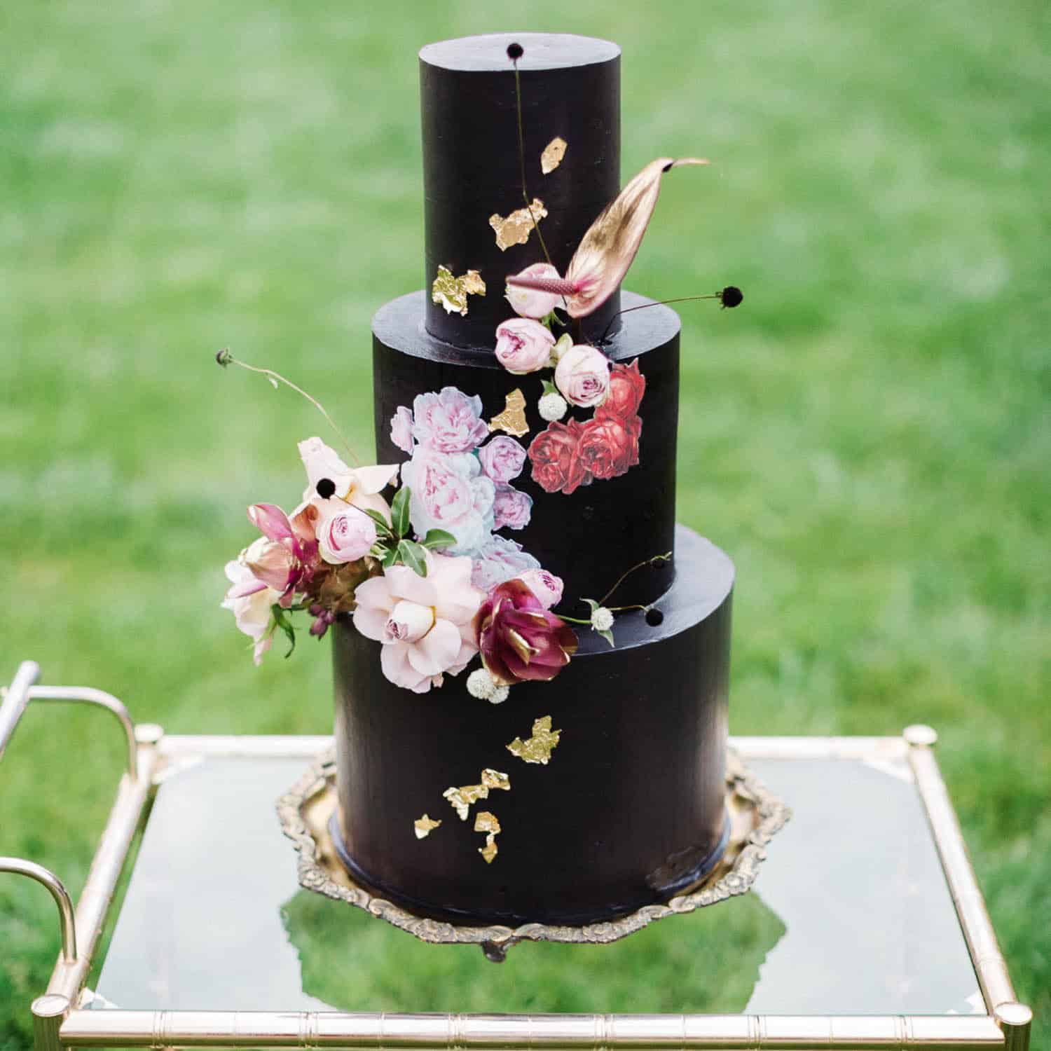 A three-tiered black cake decorated with gold leaf, painted flowers, and fresh floral accents, displayed on a glass and gold tray outdoors.