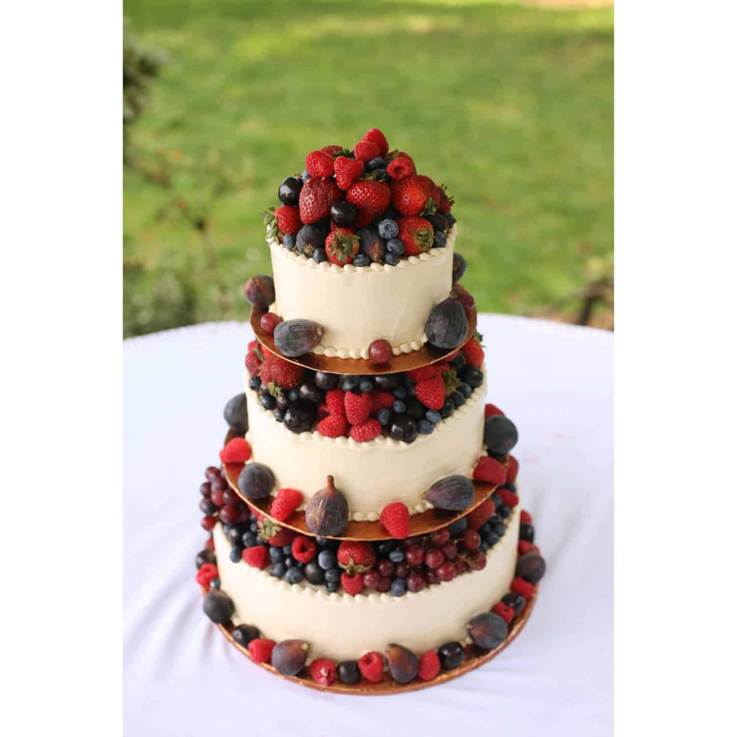 A three-tiered white frosted cake decorated with fresh strawberries, raspberries, blueberries, blackberries, and figs, displayed on a white tablecloth outdoors.