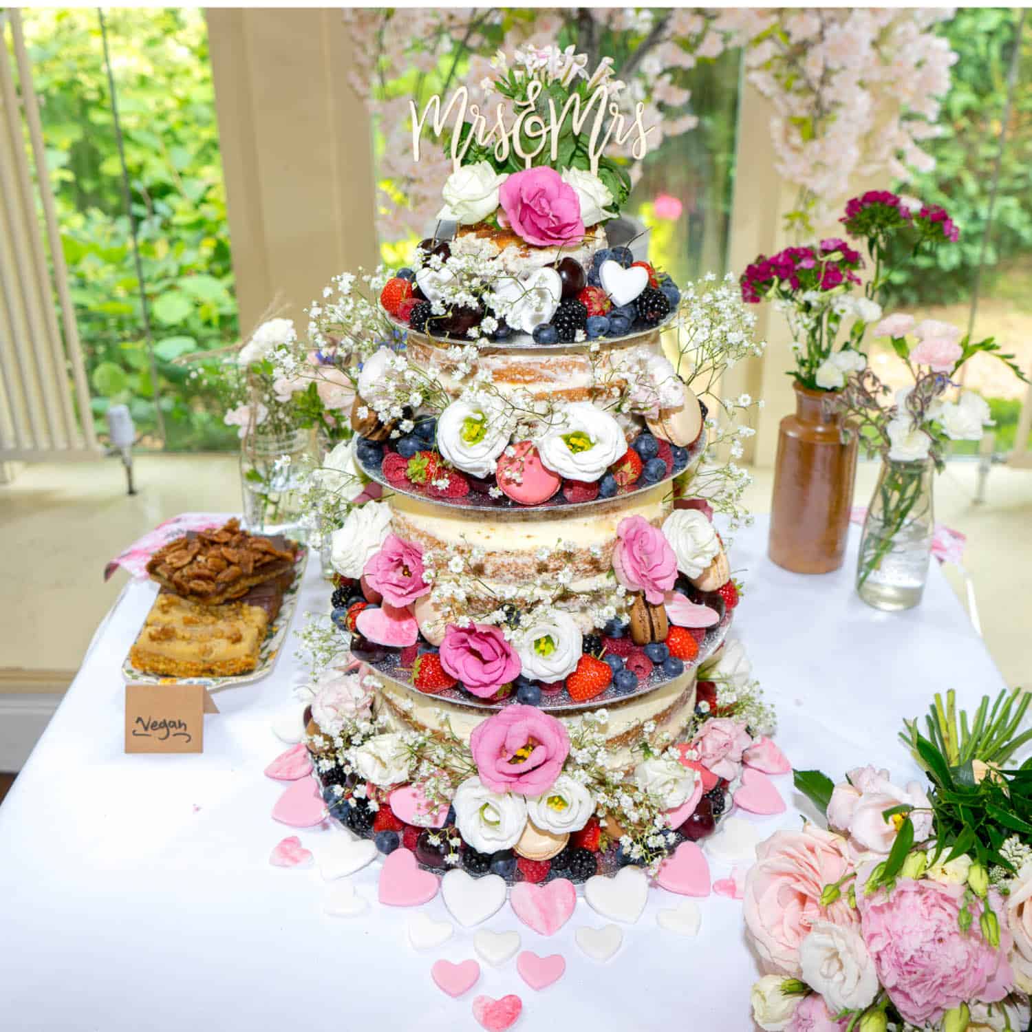 Three-tiered wedding cake decorated with fresh flowers, berries, and a "Mr and Mrs" topper sits on a table with vases of flowers and a small vegan cake nearby.