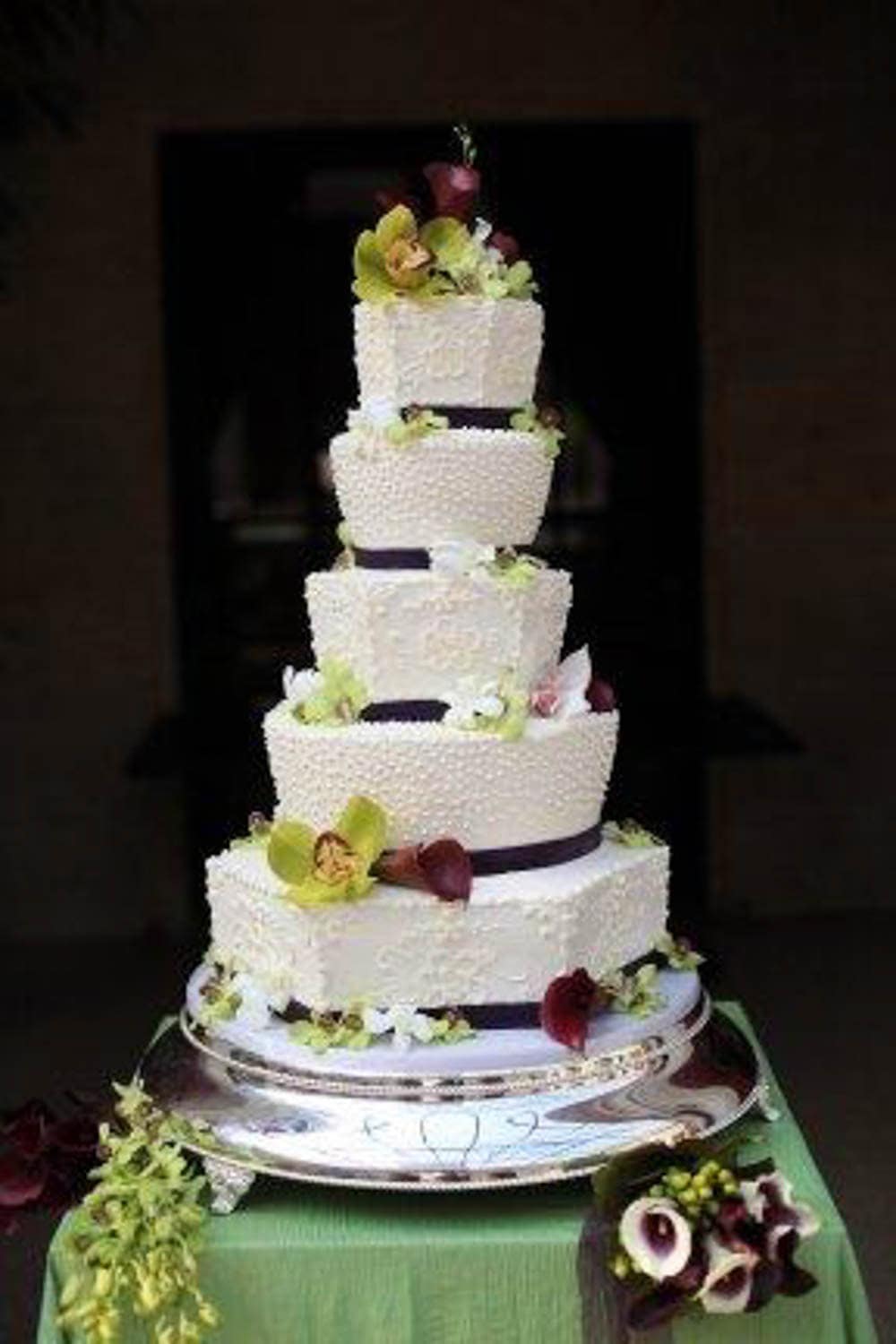 Five-tiered white wedding cake with textured icing, decorated with green and burgundy flowers, sits on a silver stand over a green cloth.