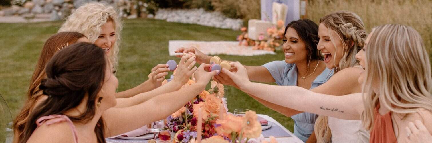 Six women sitting outdoors around a decorated table, smiling and holding up colorful macarons in a celebratory gesture.