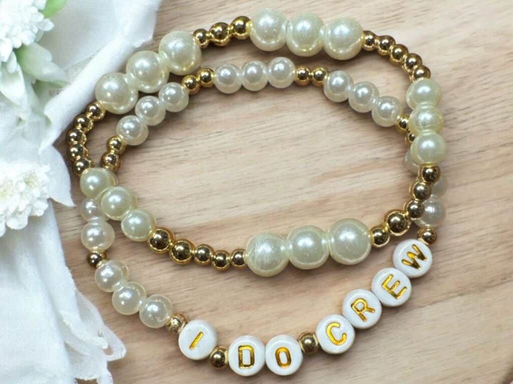 Two handcrafted beaded bracelets with pearl and gold beads, one featuring white letter beads spelling “I DO CREW,” displayed on a wooden surface next to white floral lace.