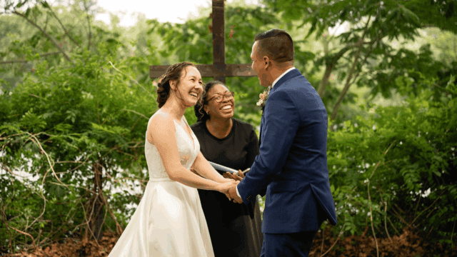 Fallon of About Love Celebrations officiating an outdoor wedding ceremony for Kina and Josh in Cumberland County, NJ. All three are laughing joyfully under a wooden cross, surrounded by lush greenery.