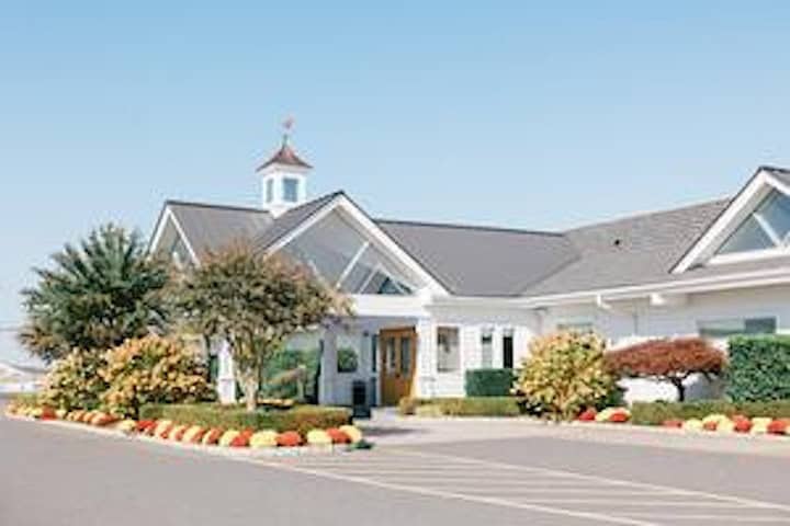 A daytime exterior shot of the Crystal Point Yacht Club, a white building with a gray roof, surrounded by manicured landscaping and a parking lot.