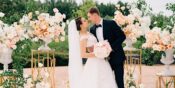 A bride and groom are captured in a loving embrace amidst elegant peach and white floral arrangements at an outdoor wedding ceremony.