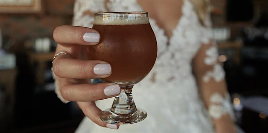 A close-up of a hand with manicured nails holding a small glass of amber beer in front of a person wearing a white lace wedding gown.