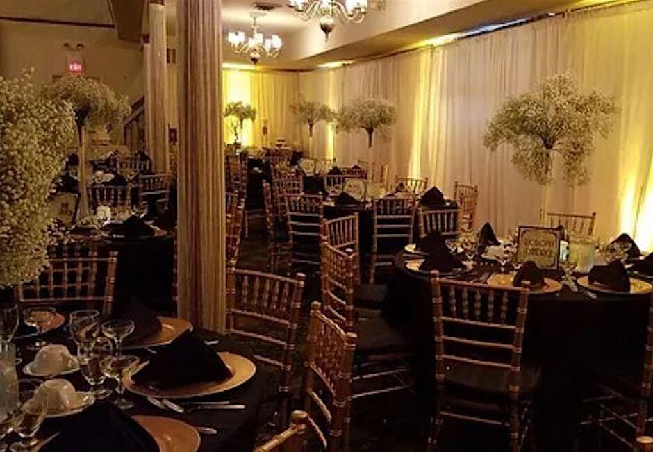 An elegant banquet hall interior decorated with round tables set with black linens and gold chairs, featuring tall floral centerpieces with baby's breath.