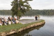 A couple exchanges vows by a serene lakeside surrounded by guests, with lush greenery and calm waters creating a romantic outdoor setting.