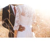 A close-up, sunlit photo of a bride wearing a lace-sleeved dress and a groom in a suit with his arm around her waist, standing in a field of tall grass.