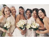 A bride smiling with her bridesmaids and family members, all holding white floral bouquets and standing closely together.