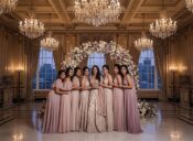 A bride in a lavish gold and champagne gown surrounded by six bridesmaids in varying shades of pink and mauve, standing under a floral arch in a grand ballroom with crystal chandeliers.
