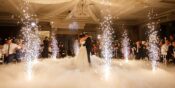 A bride and groom share a first dance surrounded by indoor sparklers and low-lying fog as guests watch in an elegant, chandelier-lit ballroom.