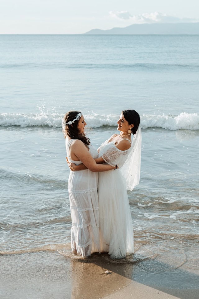 Two brides wearing lightweight white bridal gowns standing in the ocean during a beach wedding, showcasing flowy fabrics and romantic boho styles perfect for a coastal ceremony.