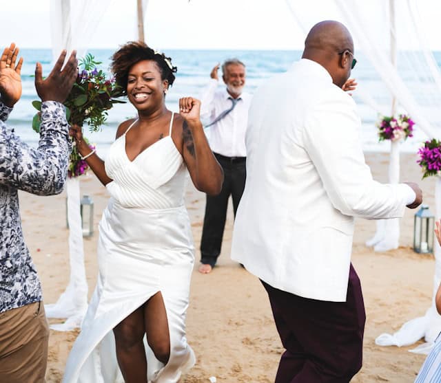 A bride in a sleek white bridal gown with a thigh slit dances joyfully on the beach during her wedding ceremony, surrounded by guests and ocean views — a perfect example of a bridal gown for a beach wedding along the Jersey Shore.