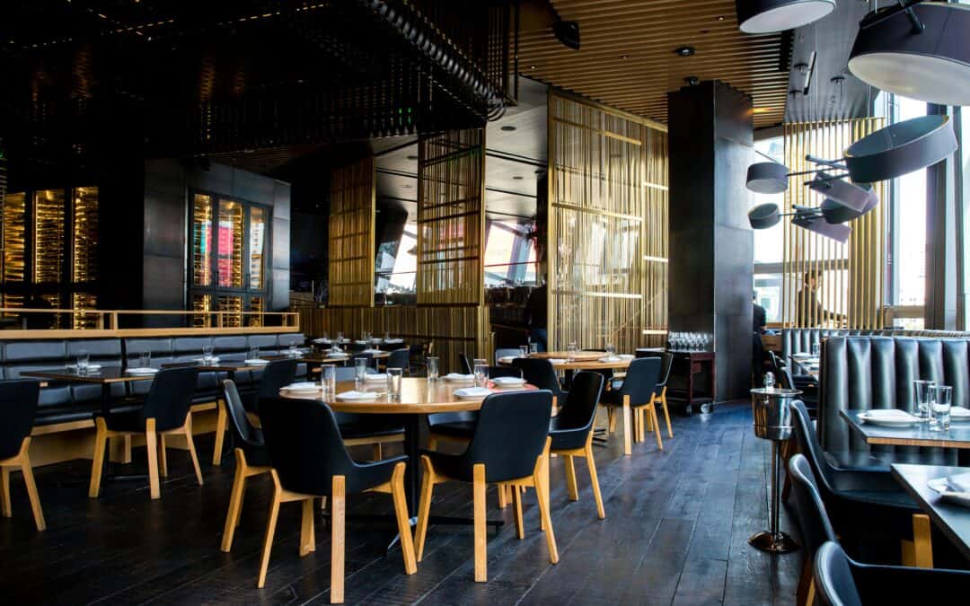 A modern upscale restaurant with round wooden tables, black chairs, gold accent dividers, and soft ambient lighting. The room is empty and fully set for dining.