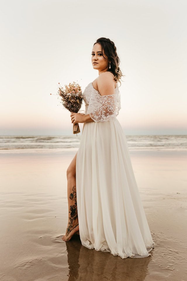 Tattooed bride in an off-the-shoulder lace bridal gown for a beach wedding, holding a bouquet while standing barefoot on the shoreline at sunset.