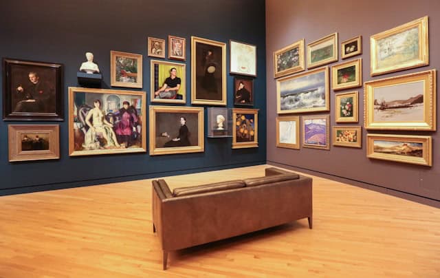 A quiet art gallery room with wooden floors, dark-toned walls, and a salon-style display of framed artwork surrounding a central leather bench.