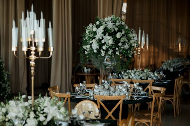 Elegant wedding reception table with white floral centerpiece, candles, and wooden chairs in a warmly lit venue.